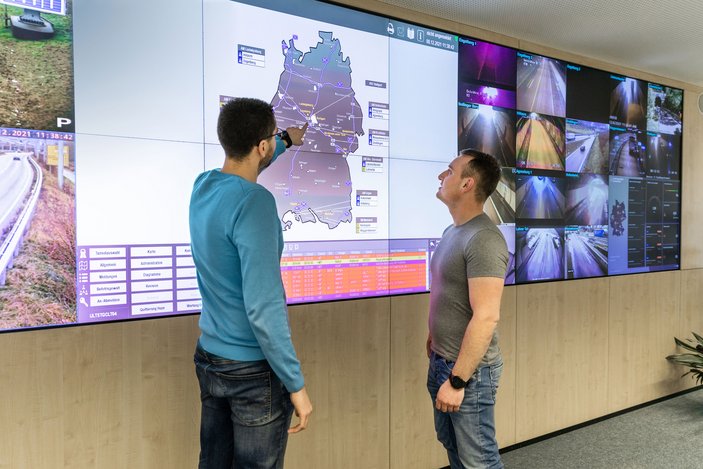 Zwei Männer betrachten eine große digitale Anzeigetafel mit Verkehrsinformationen.
