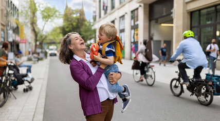 Ältere Frau hält lachend ein Kind in einer belebten Innenstadt hoch.