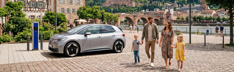 Eine Familie spaziert auf einem Kopfsteinpflasterplatz in der Altstadt von Heidelberg, während ein silbernes Elektroauto an einer Ladestation geparkt ist. Im Hintergrund sind die Alte Brücke und das Heidelberger Schloss zu sehen.