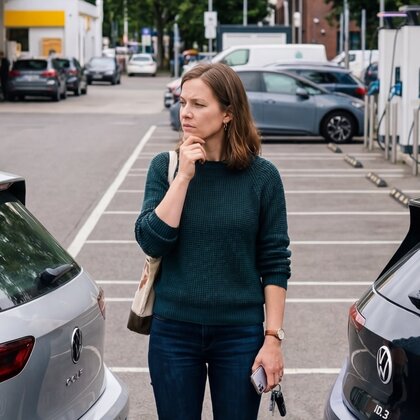 Eine nachdenkliche Frau mit Autoschlüsseln steht auf einem Parkplatz zwischen zwei Autos, im Hintergrund sind Ladestationen für Elektroautos sichtbar.