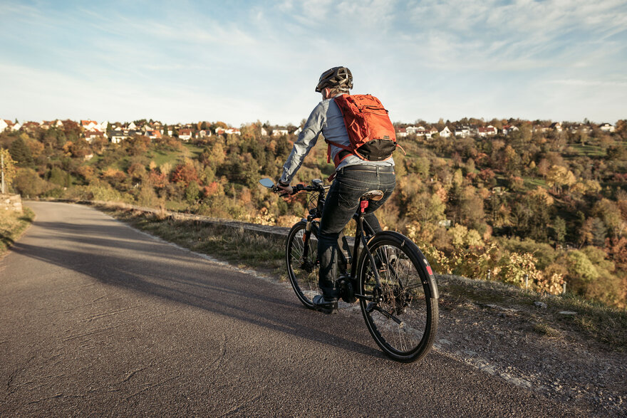 Mann mit Rucksack fährt auf einem Fahrradweg durch eine herbstliche Hügellandschaft.
