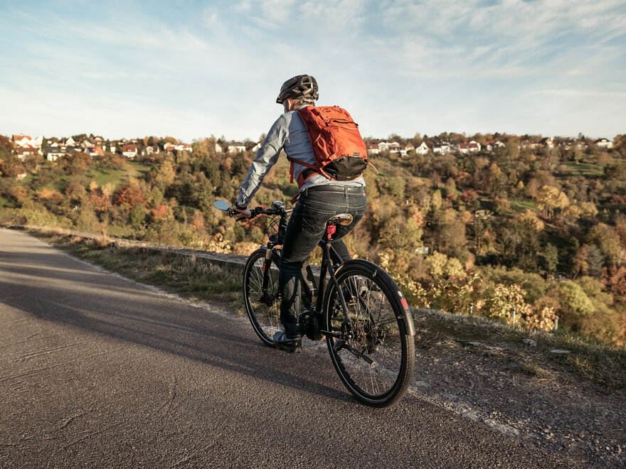 Mann mit Rucksack fährt auf einem Fahrradweg durch eine herbstliche Hügellandschaft.