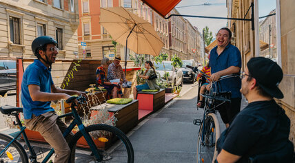 Belebte Straßenszene an einem Parklet: Ein Mann auf dem Fahrrad und ein Mann im Rollstuhl unterhalten sich lachend auf dem Gehweg vor einer Außengastronomie.