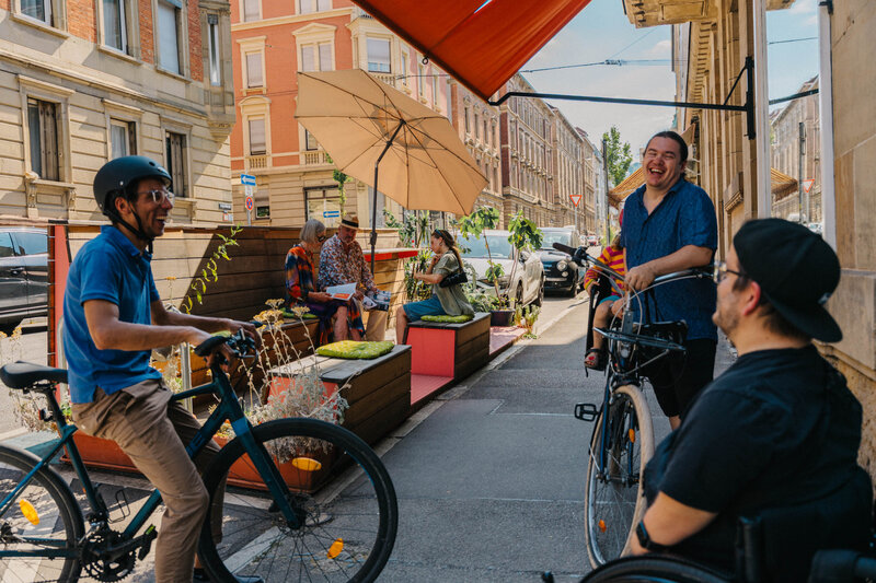 Belebte Straßenszene an einem Parklet: Ein Mann auf dem Fahrrad und ein Mann im Rollstuhl unterhalten sich lachend auf dem Gehweg vor einer Außengastronomie.