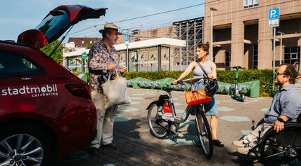 Drei Menschen im Gespräch an einer Mobilitätsstation: Ein Mann belädt den Kofferraum eines roten Carsharing-Autos von stadtmobil, eine Frau steht mit einem RegioRad-Mietfahrrad daneben und ein Mann sitzt im Rollstuhl.