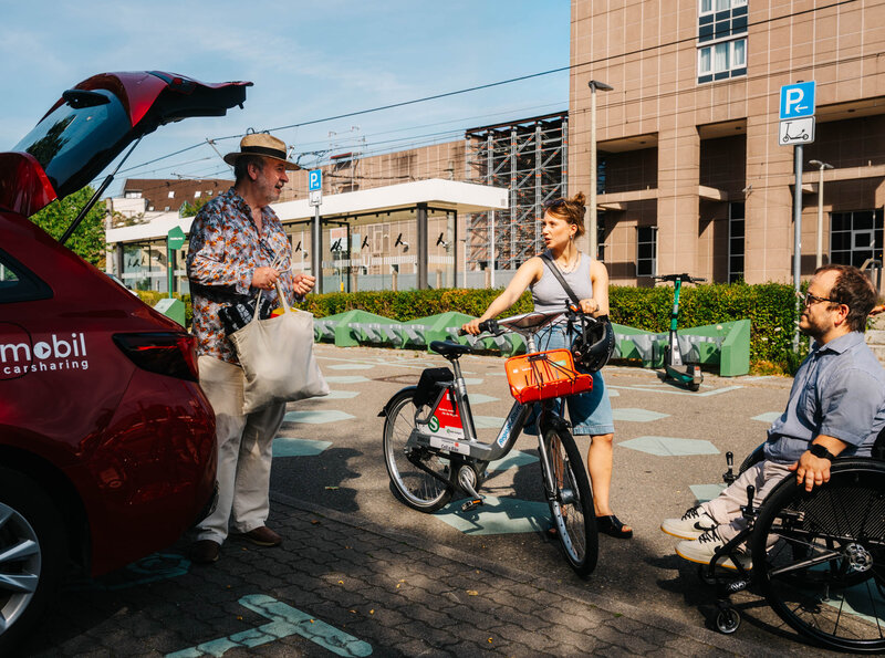Drei Menschen im Gespräch an einer Mobilitätsstation: Ein Mann belädt den Kofferraum eines roten Carsharing-Autos von stadtmobil, eine Frau steht mit einem RegioRad-Mietfahrrad daneben und ein Mann sitzt im Rollstuhl.