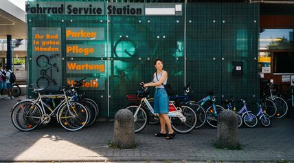 Frau schiebt ein Mietfahrrad vor der grünen Fassade einer Fahrrad-Service-Station, an der mehrere Fahrräder geparkt sind.