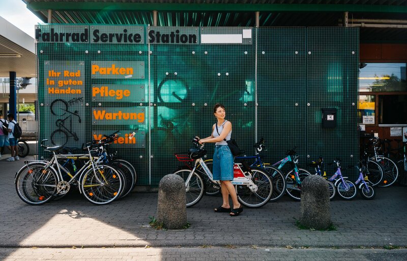 Frau schiebt ein Mietfahrrad vor der grünen Fassade einer Fahrrad-Service-Station, an der mehrere Fahrräder geparkt sind.