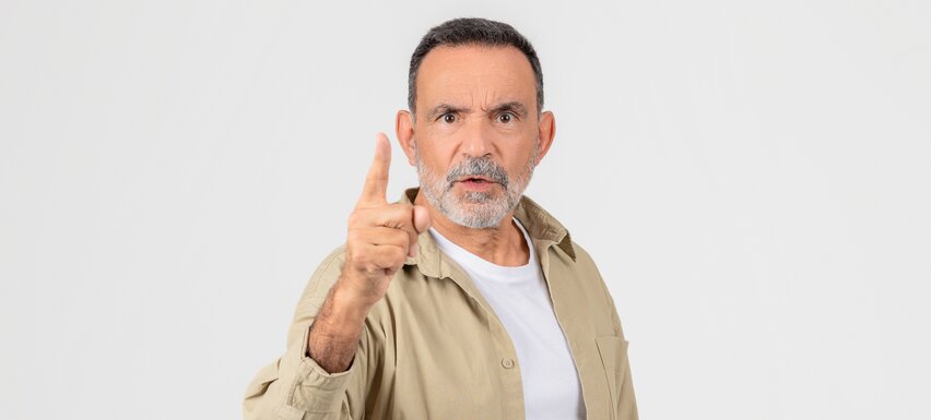 Älterer Mann mit grauem Bart und ernstem Blick hebt mahnend den Zeigefinger vor weißem Hintergrund.