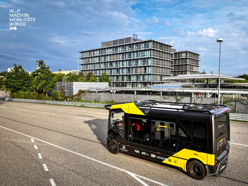 Ein kleiner Bus der sich auf einer Straße bewegt, im Hintergrund ist ein großes Gebäude.