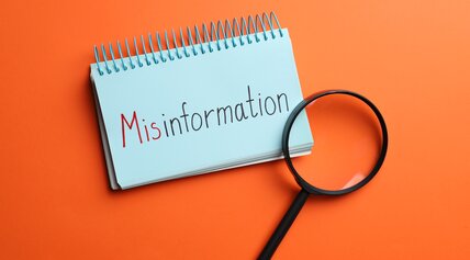 Blaues Notizbuch mit der handschriftlichen Aufschrift „Misinformation“ und eine Lupe auf leuchtend orangefarbenem Hintergrund.