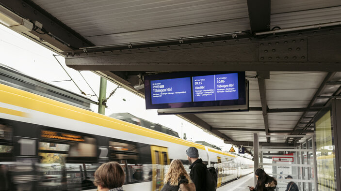 Fahrgäste warten am Bahnsteig, während ein Zug vorbeifährt; digitale Anzeigetafel.