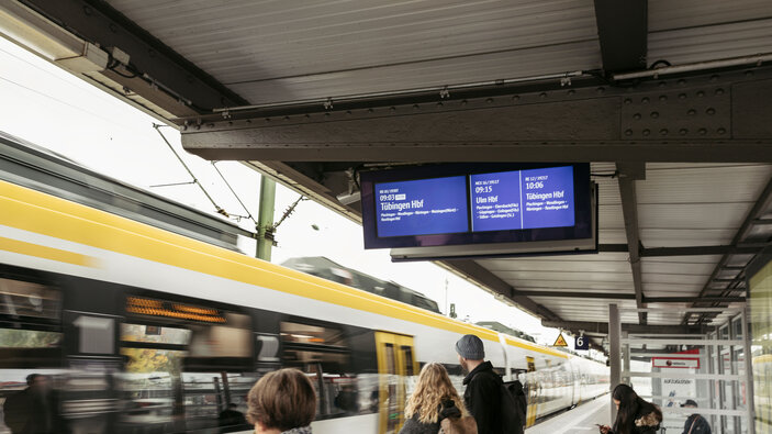 Fahrgäste warten am Bahnsteig, während ein Zug vorbeifährt; digitale Anzeigetafel.