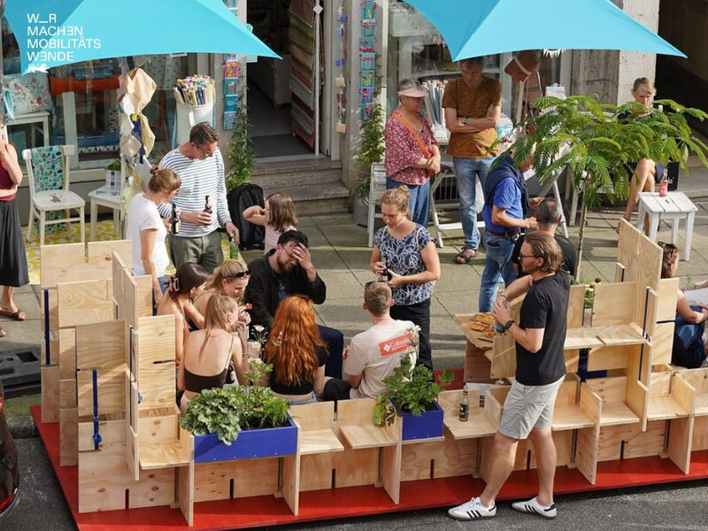 Eine große Gruppe Menschen die auf Sitzmöglichkeiten aus Holz vor einem Haus sitzen und sich unterhalten.