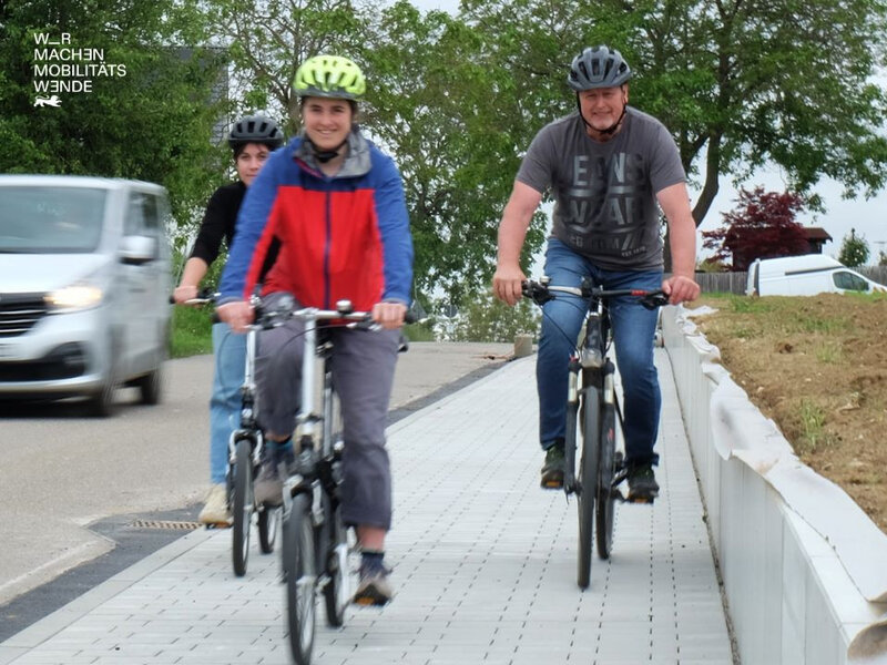 Drei Personen auf unterschiedlichen Fahrrädern die neben der Straße fahren. Ein Auto im Hintergrund.