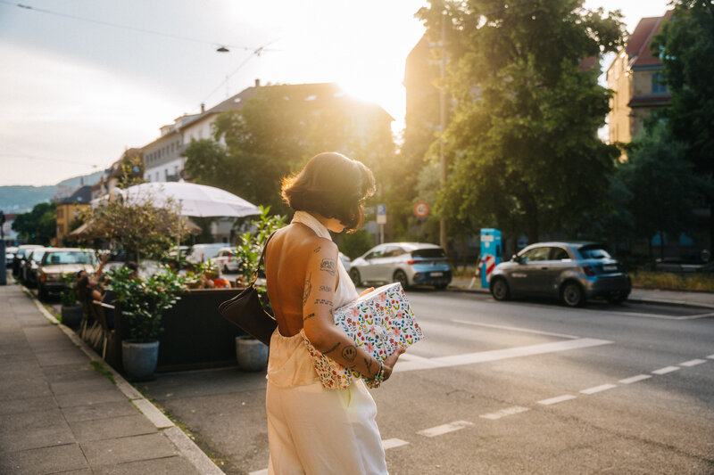  Frau im rückenfreien weißen Outfit trägt ein bunt verpacktes Geschenk bei warmem Abendlicht entlang eines Gehwegs.