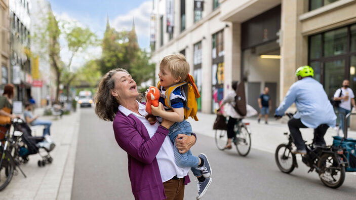 Ältere Frau hält lachend ein Kind in einer belebten Innenstadt hoch.