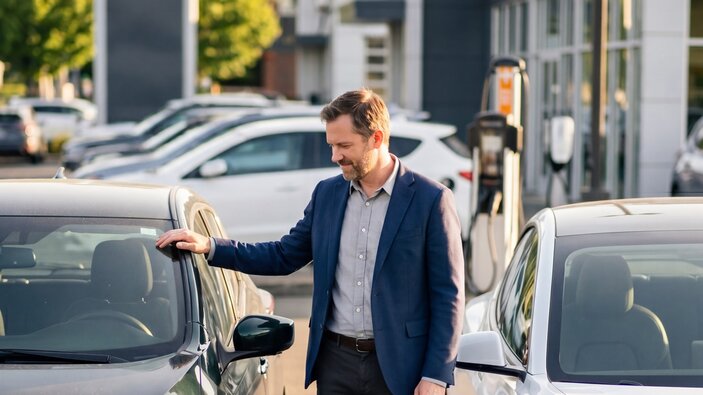 Ein Mann im Anzug steht auf einem Hof eines Autohändlers zwischen einem weisen und schwarzen Auto.