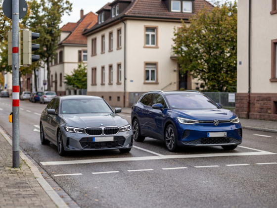 Ein grauer BMW 3er und ein blauer Volkswagen ID.4 stehen nebeneinander an einer Ampel auf einer Straße. Im Hintergrund sind Wohngebäude und Bäume zu sehen.