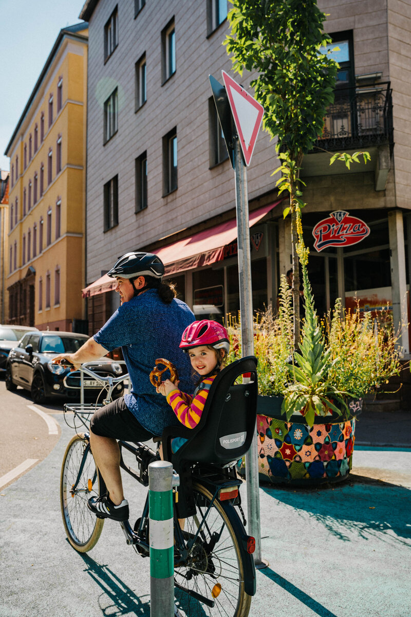 Ein Mann fährt Fahrrad mit einer kleinen Tochter im Kindersitz, die eine Laugenbrezel hält, auf einer blau markierten Spur vorbei an einer Pizzeria.