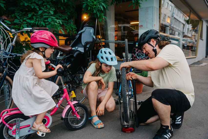 Ein kniender Mann pumpt auf dem Gehweg den Reifen eines Kinderfahrrads auf, während zwei Mädchen mit Fahrradhelmen aufmerksam zusehen.