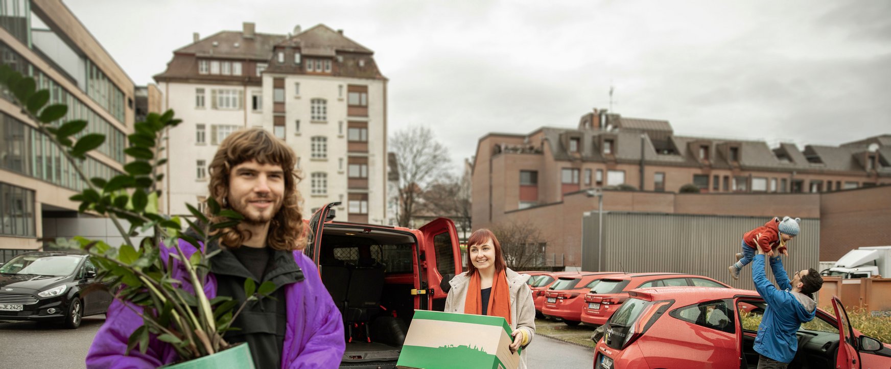 Zwei Personen laden Gegenstände aus einem roten Carsharing-Auto, Familie im Hintergrund.