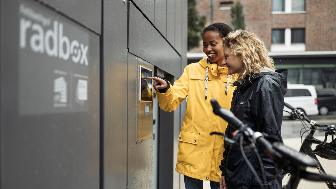 Zwei Frauen bedienen einen Bildschirm an einer Radbox Fahrradstation.