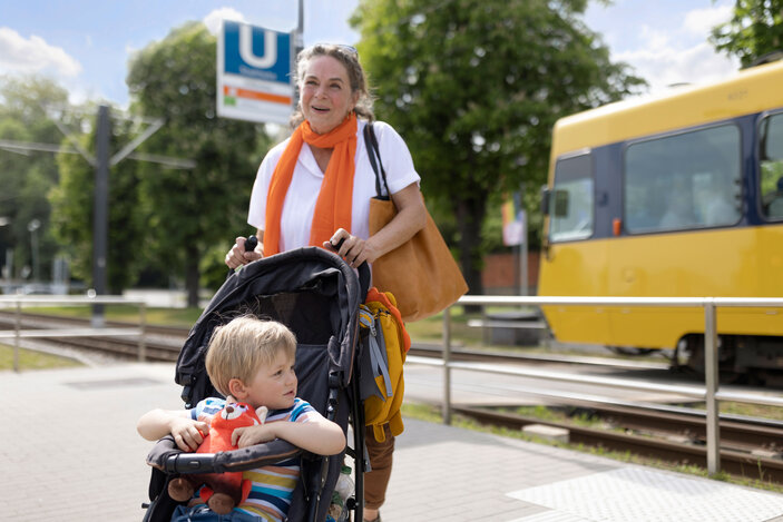 Frau schiebt Kinderwagen mit Kind an einer Straßenbahnhaltestelle, gelbe Bahn fährt vorbei.