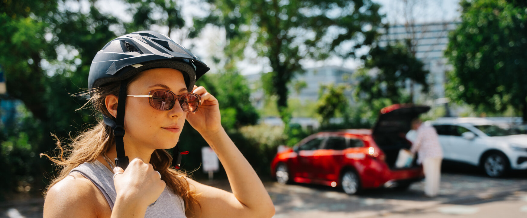 Nahaufnahme einer jungen Frau mit Fahrradhelm und Sonnenbrille auf einem Parkplatz, während im Hintergrund ein roter Kombi beladen wird.
