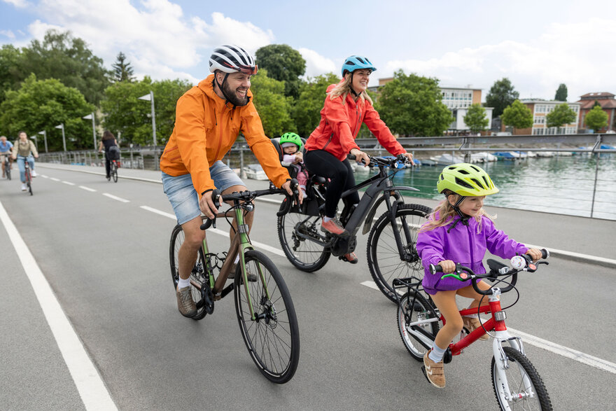 Familie mit zwei Kindern fährt lachend auf Fahrrädern über eine Brücke am Wasser.