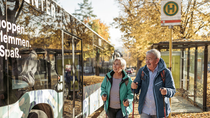 Älteres Paar mit Wanderstöcken geht im Herbst zu einem Bus an einer Haltestelle.