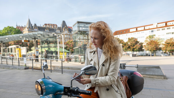 Frau sitzt auf einem blauen E-Roller in der Stadt und nutzt ihr Smartphone