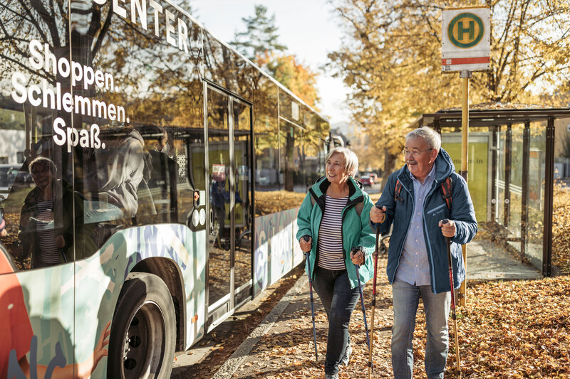 Älteres Paar mit Wanderstöcken geht im Herbst zu einem Bus an einer Haltestelle.