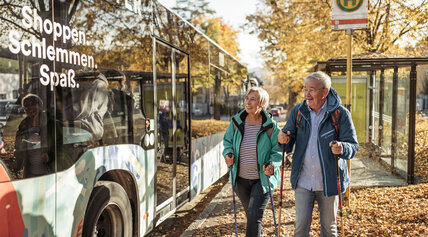 Älteres Paar mit Wanderstöcken geht im Herbst zu einem Bus an einer Haltestelle.