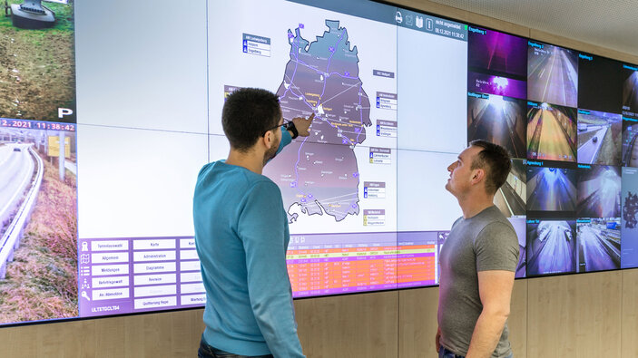 Zwei Männer betrachten eine große digitale Anzeigetafel mit Verkehrsinformationen.