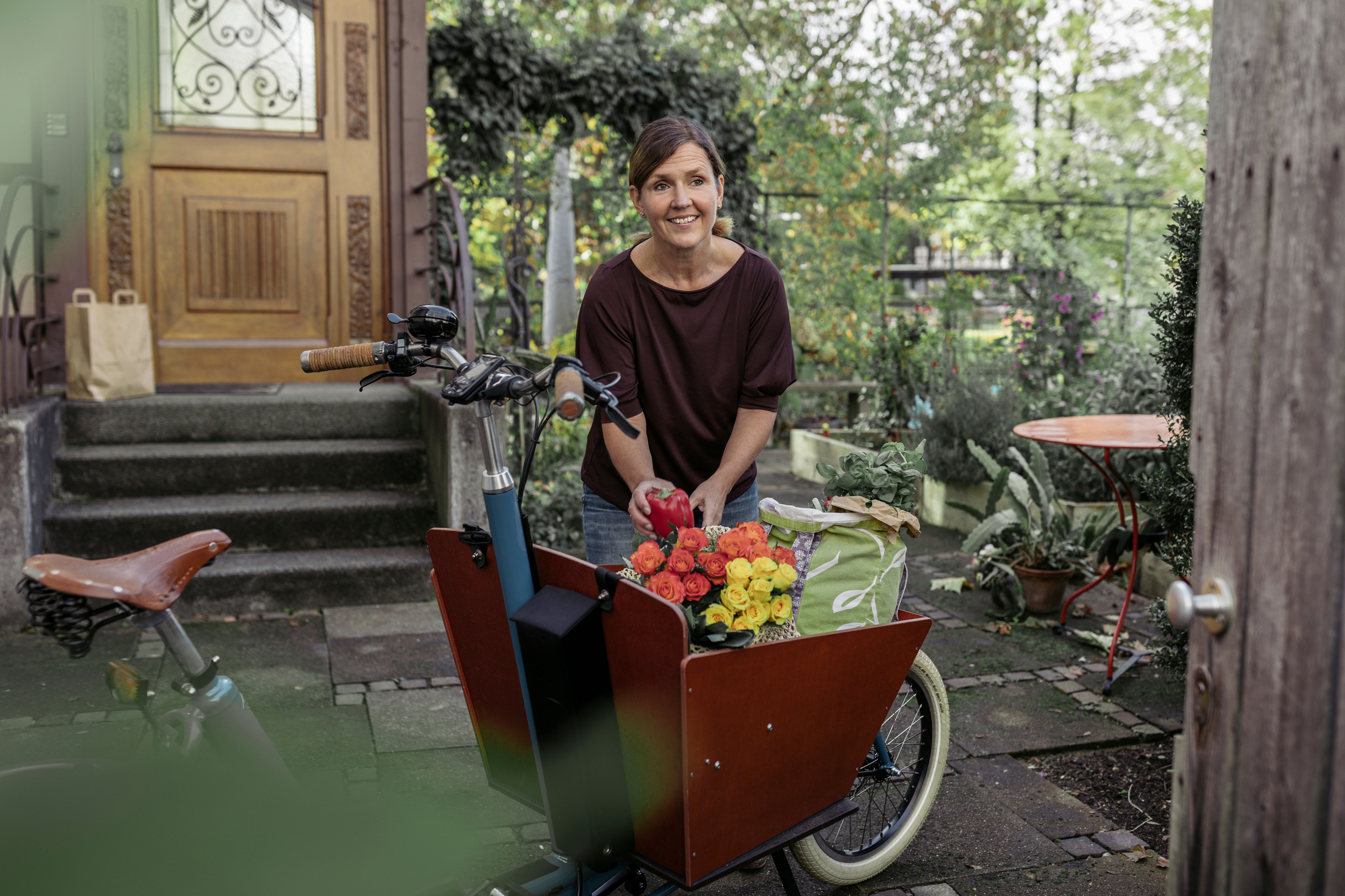 Lächelnde Frau steht mit Einkauf und Blumen vor einem Lastenrad vor ihrem Haus.