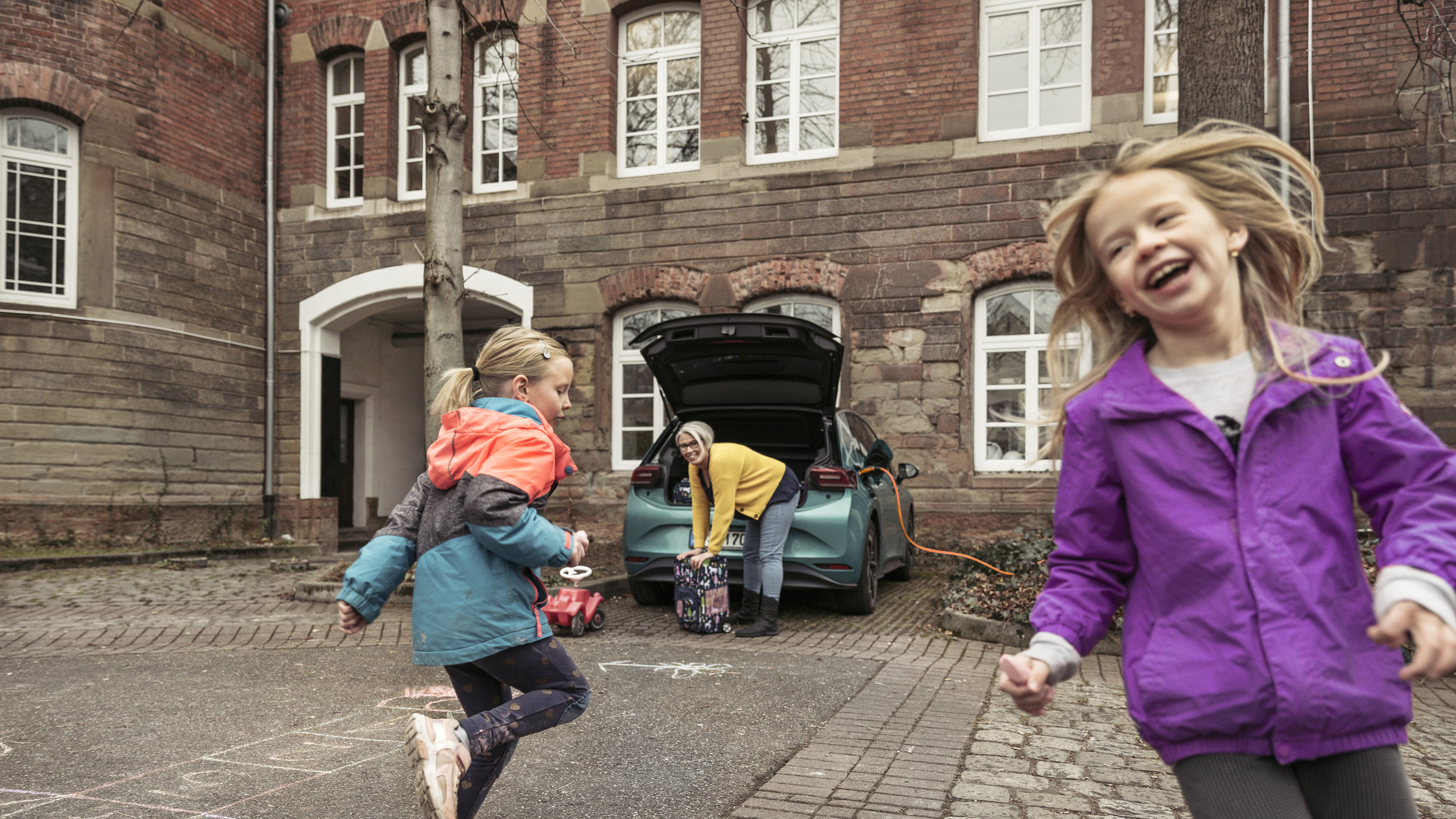 Kinder spielen vor einem E-Auto, das geladen wird, während eine Frau den Kofferraum belädt.