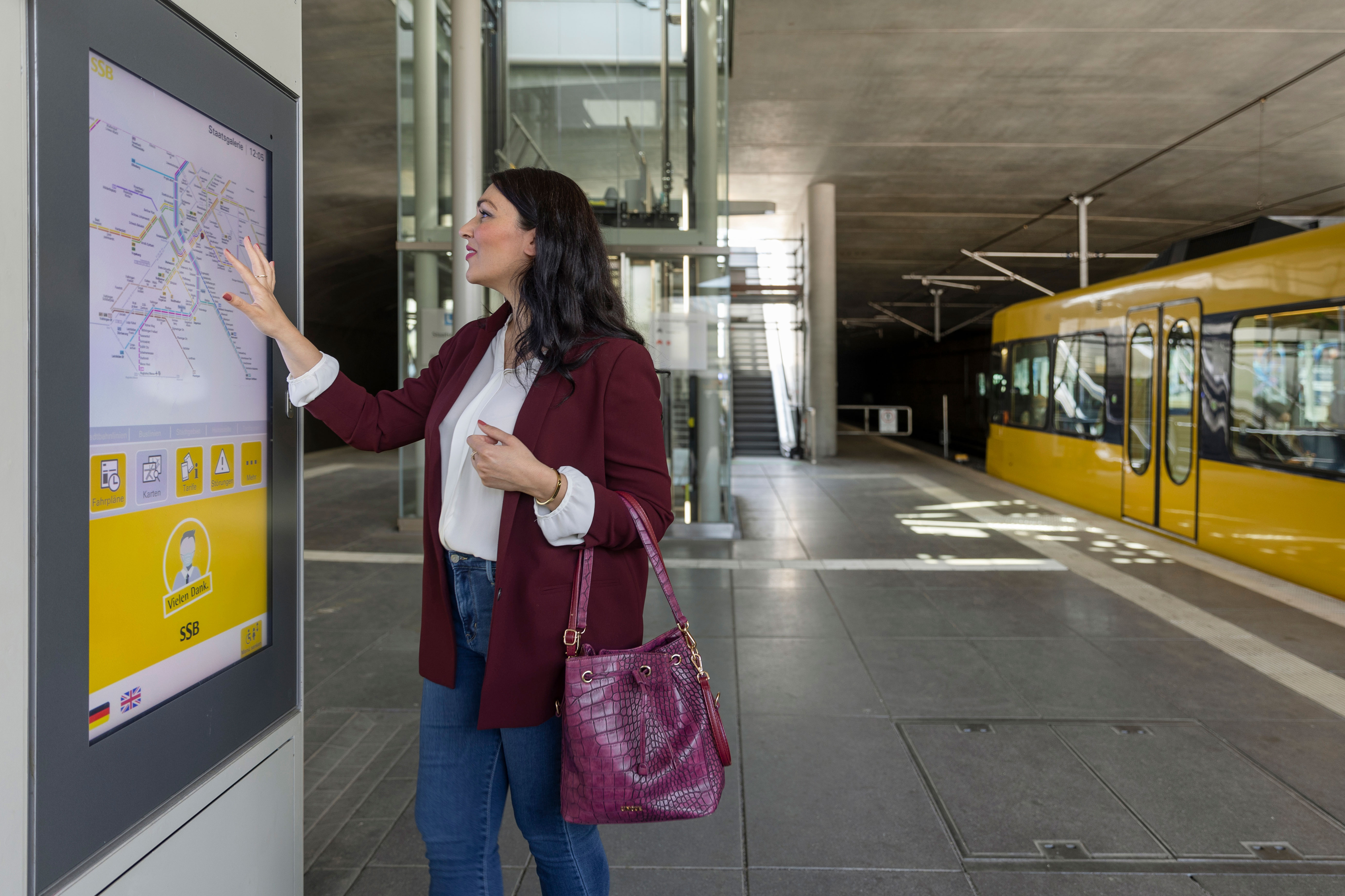 Frau nutzt digitalen Informationsbildschirm mit Fahrplan an einer Stadtbahnhaltestelle.