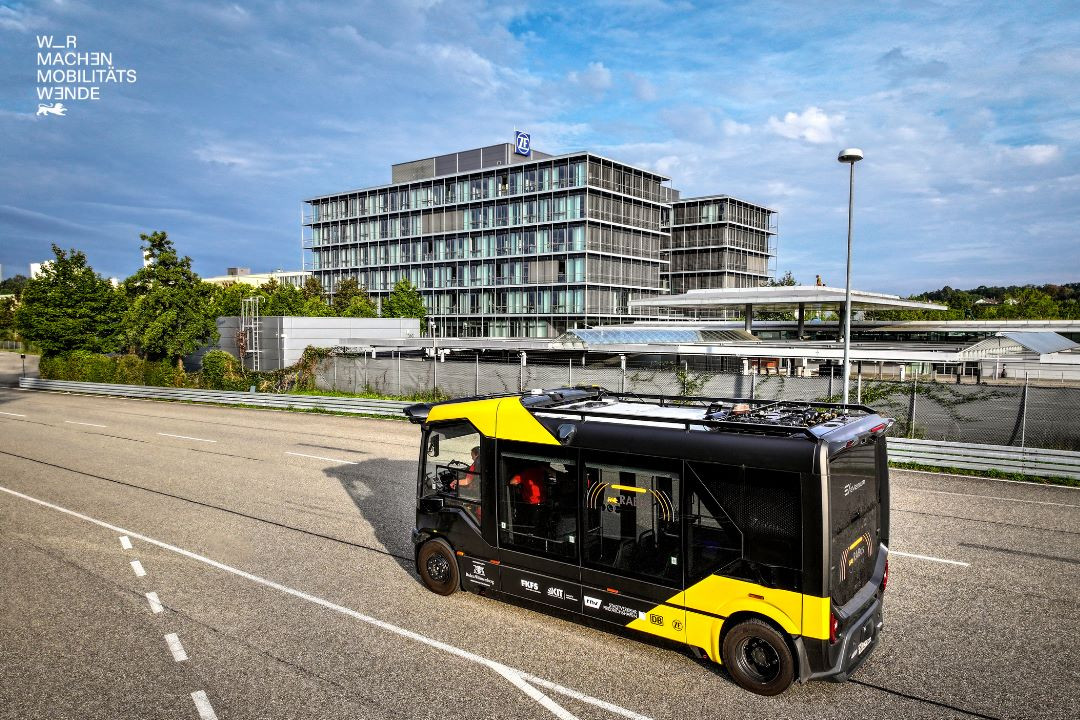 Ein kleiner Bus der sich auf einer Straße bewegt, im Hintergrund ist ein großes Gebäude.