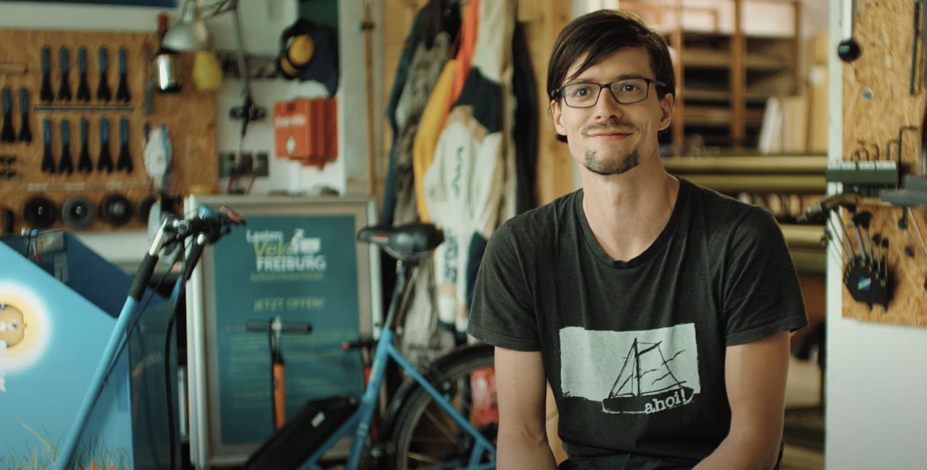 Ein Mann mit Brille, welcher in einer Garage sitzt, im Hintergrund ist ein Fahrrad.