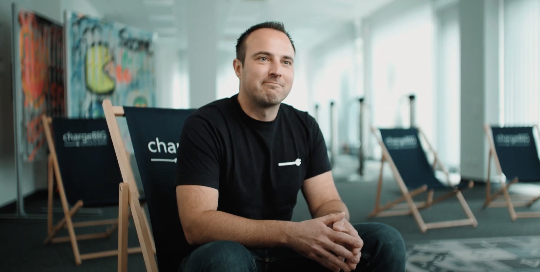 Ein Mann in schwarzem T-Shirt den in einem Liegestuhl in einem Innenraum sitzt, weitere Stühle im Hintergrund.