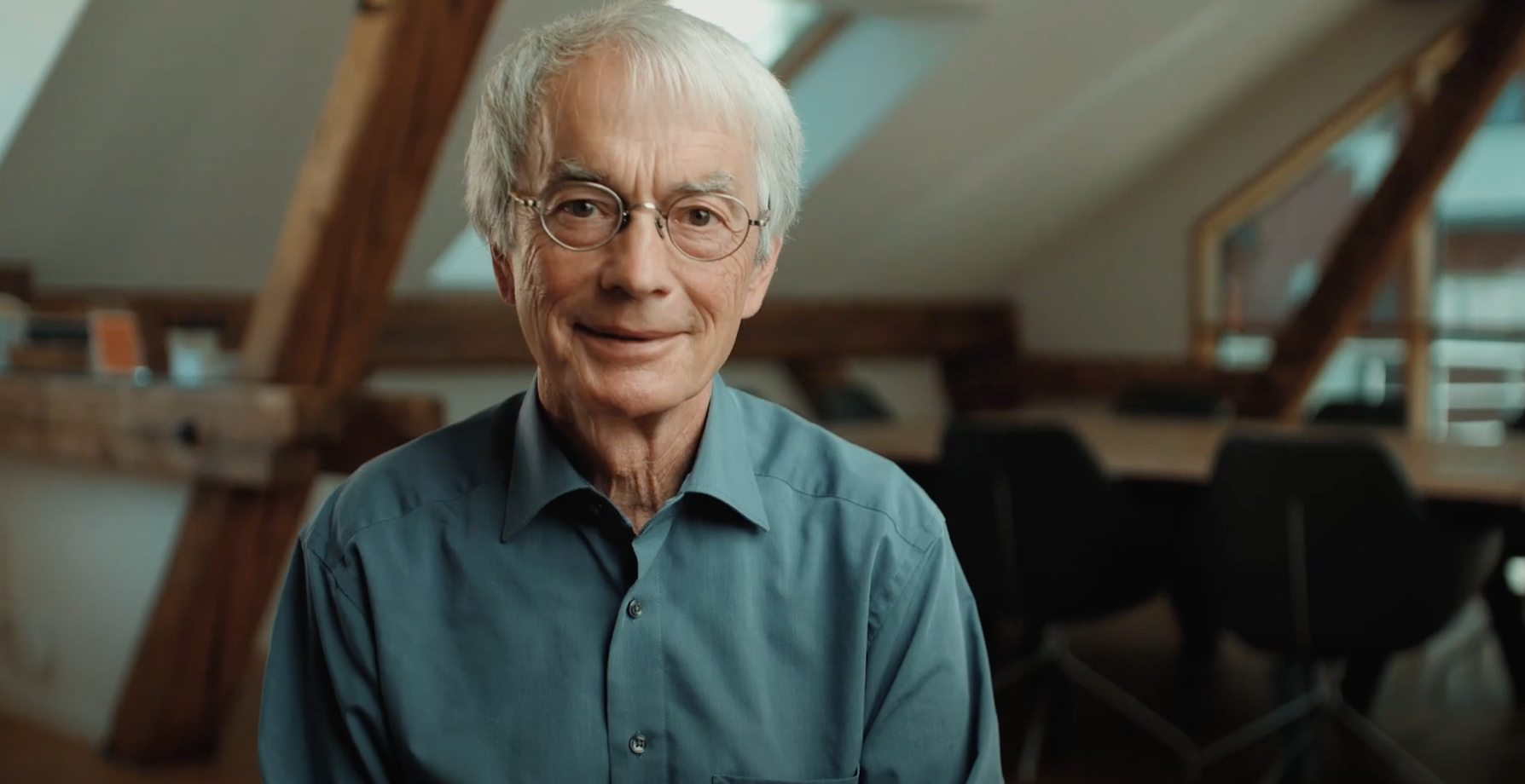 Ein älterer Mann sitzt in einer Wohnung. Wohnraum mit Tischen und Stühlen im Hintergrund. 