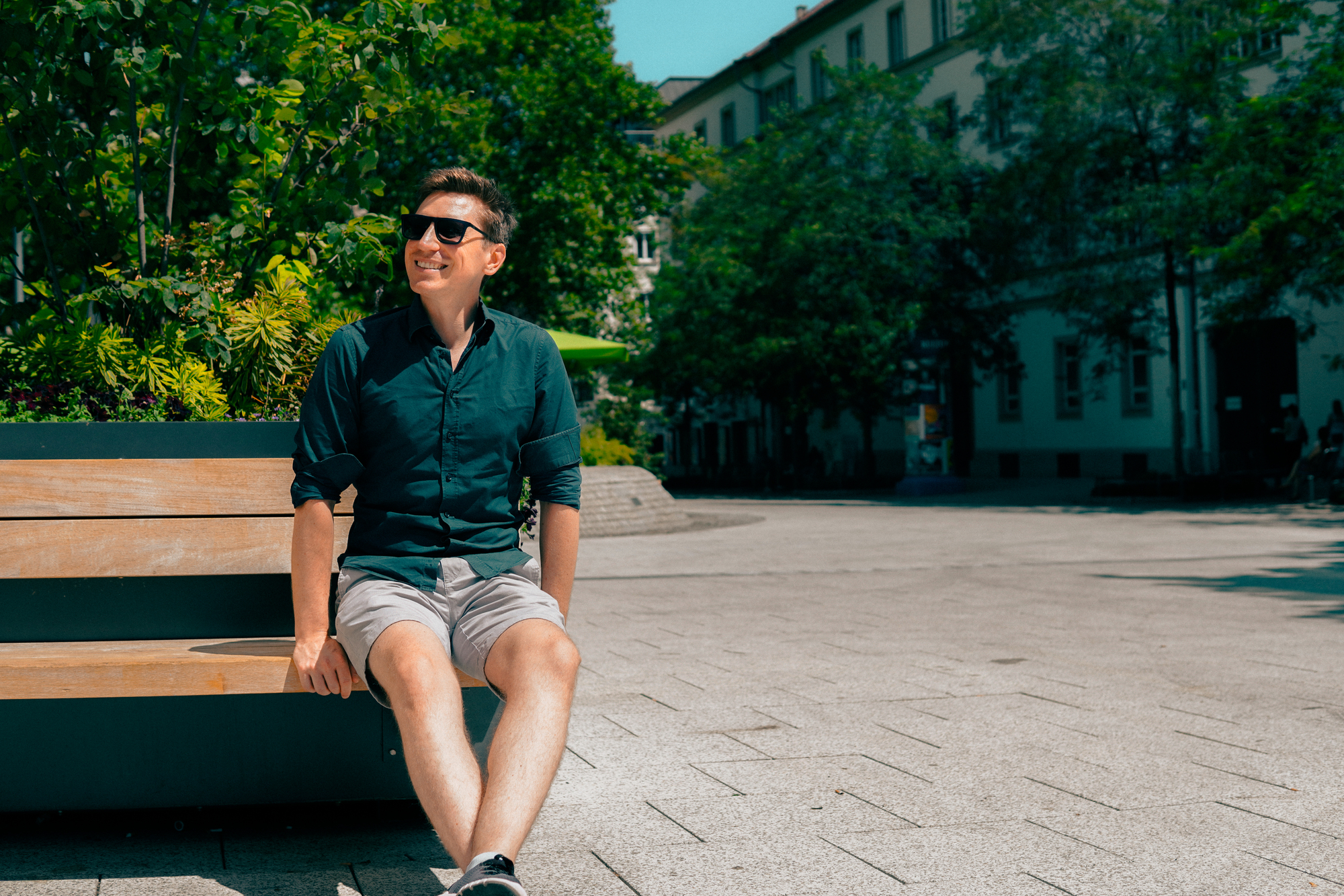 Ein Mann mit Sonnenbrille sitzt auf einer Bank auf einem großen Platz und genießt das Wetter.
