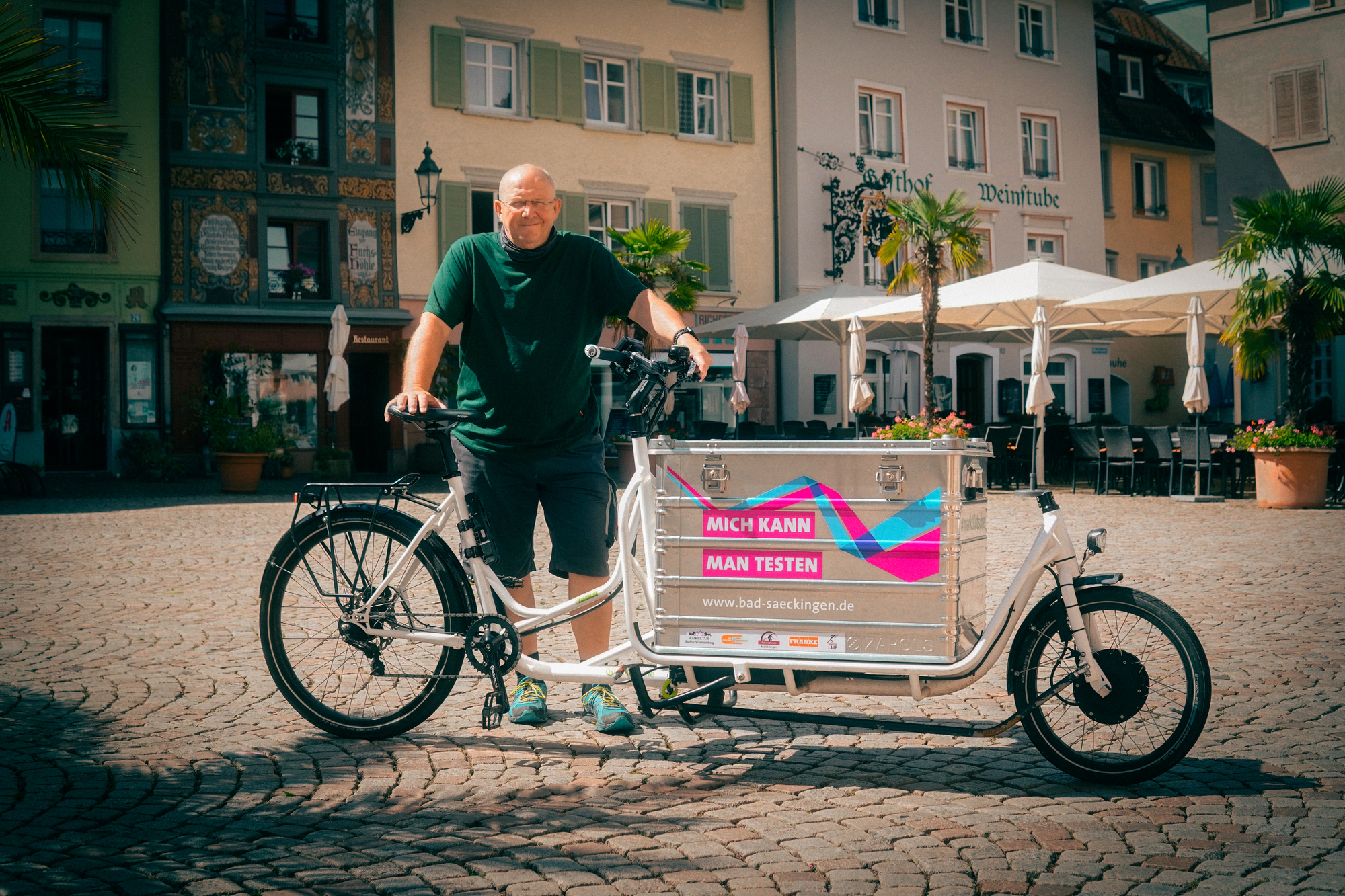 Ein Mann steht mit seinem Lastenrad auf einem Platz in einer Innenstadt.