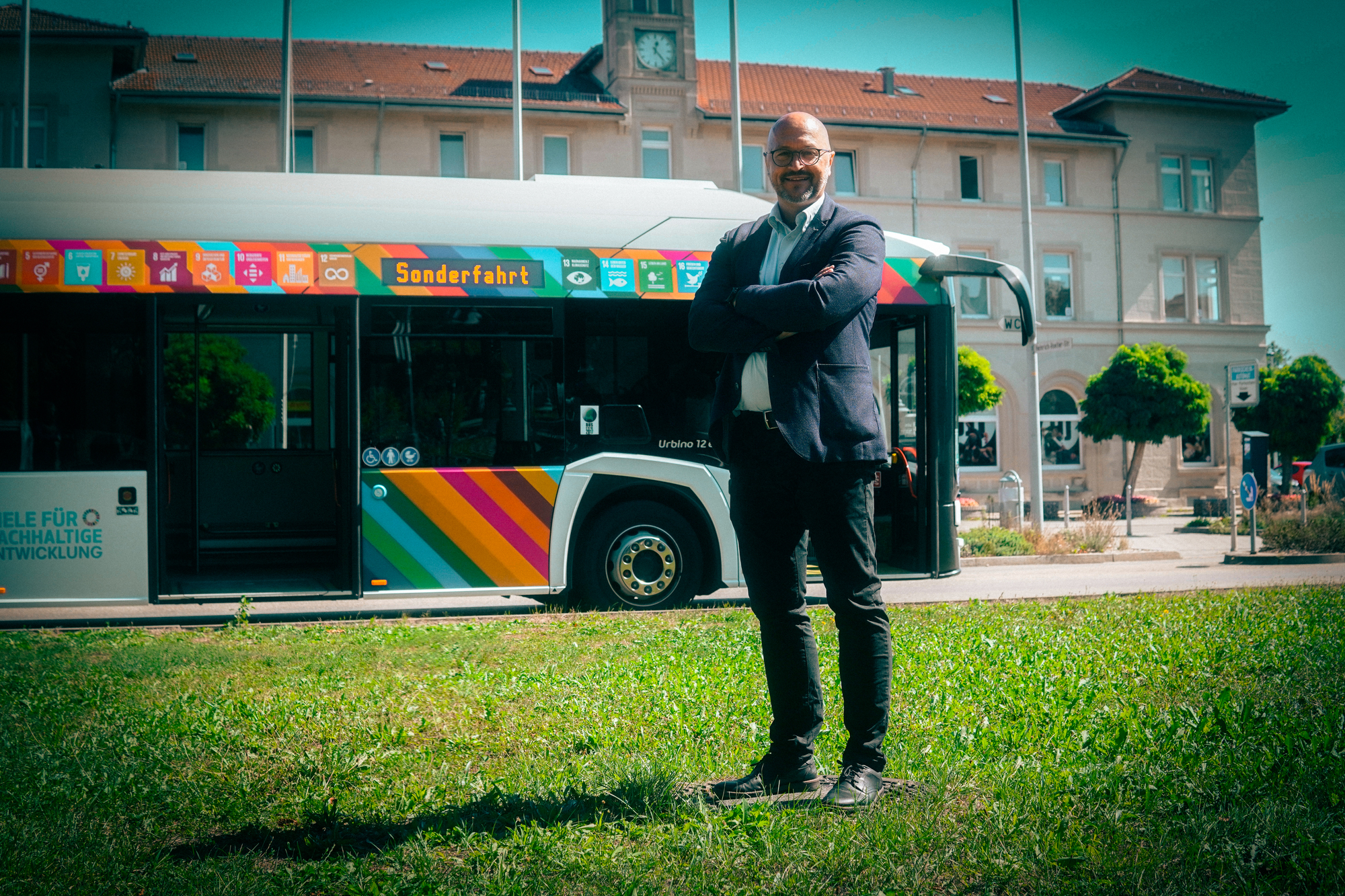 Ein Mann mit verschränkten Armen, der auf einer Wiese steht, im Hintergrund steht ein Bus.