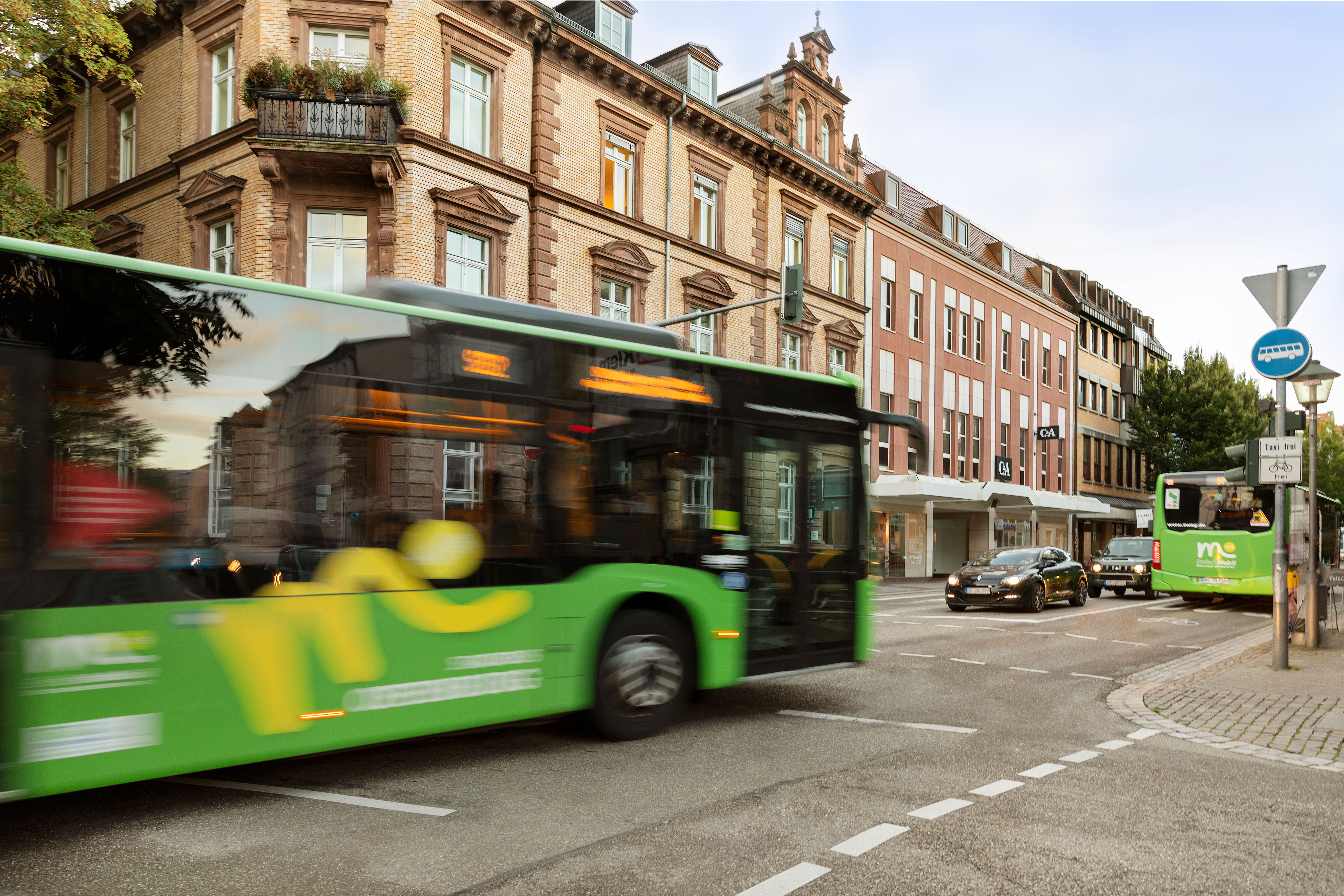 Grüner Bus fährt schnell auf einer Stadtstraße, andere Fahrzeuge im Hintergrund.