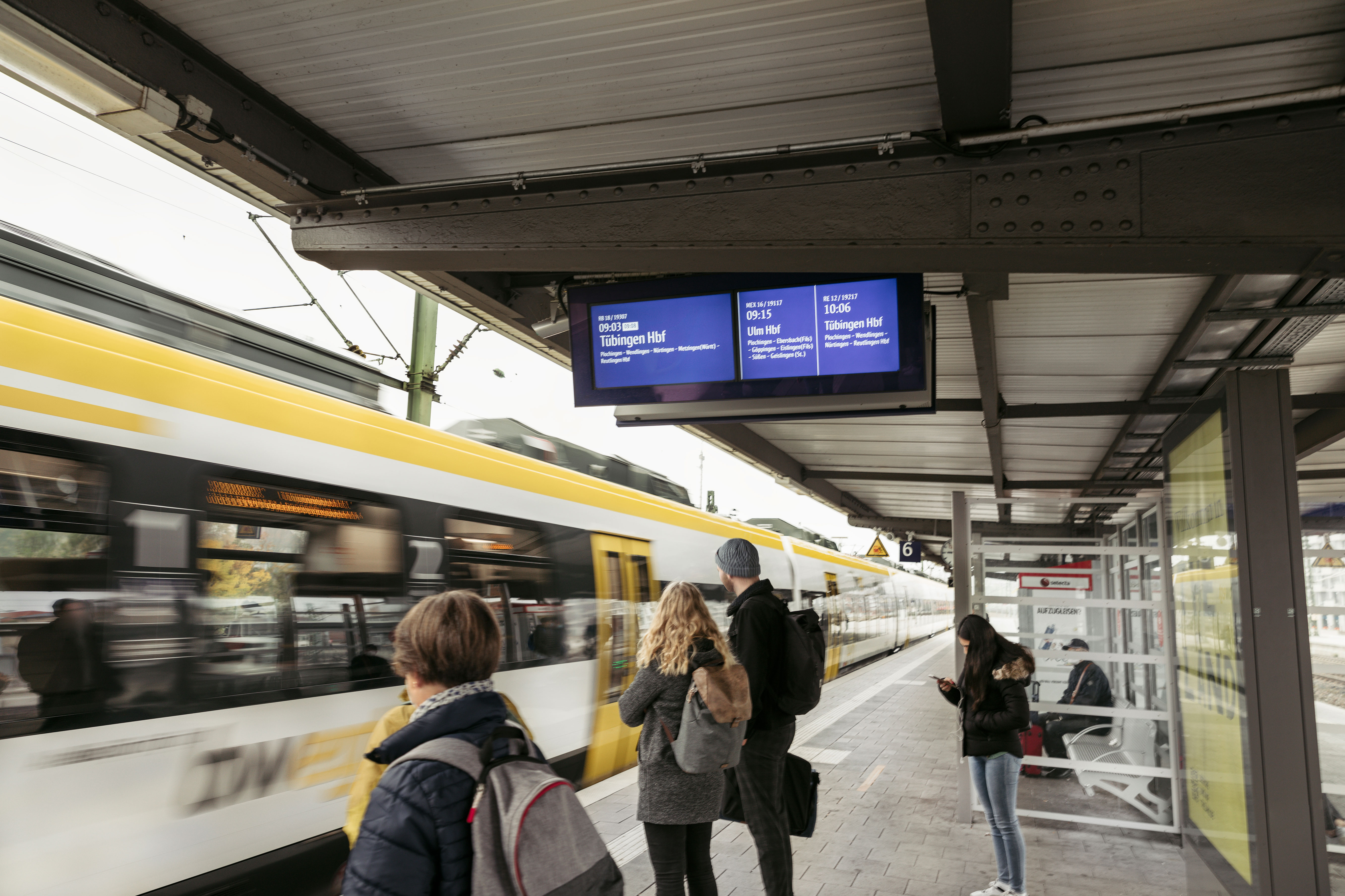 Fahrgäste warten am Bahnsteig, während ein Zug vorbeifährt; digitale Anzeigetafel.