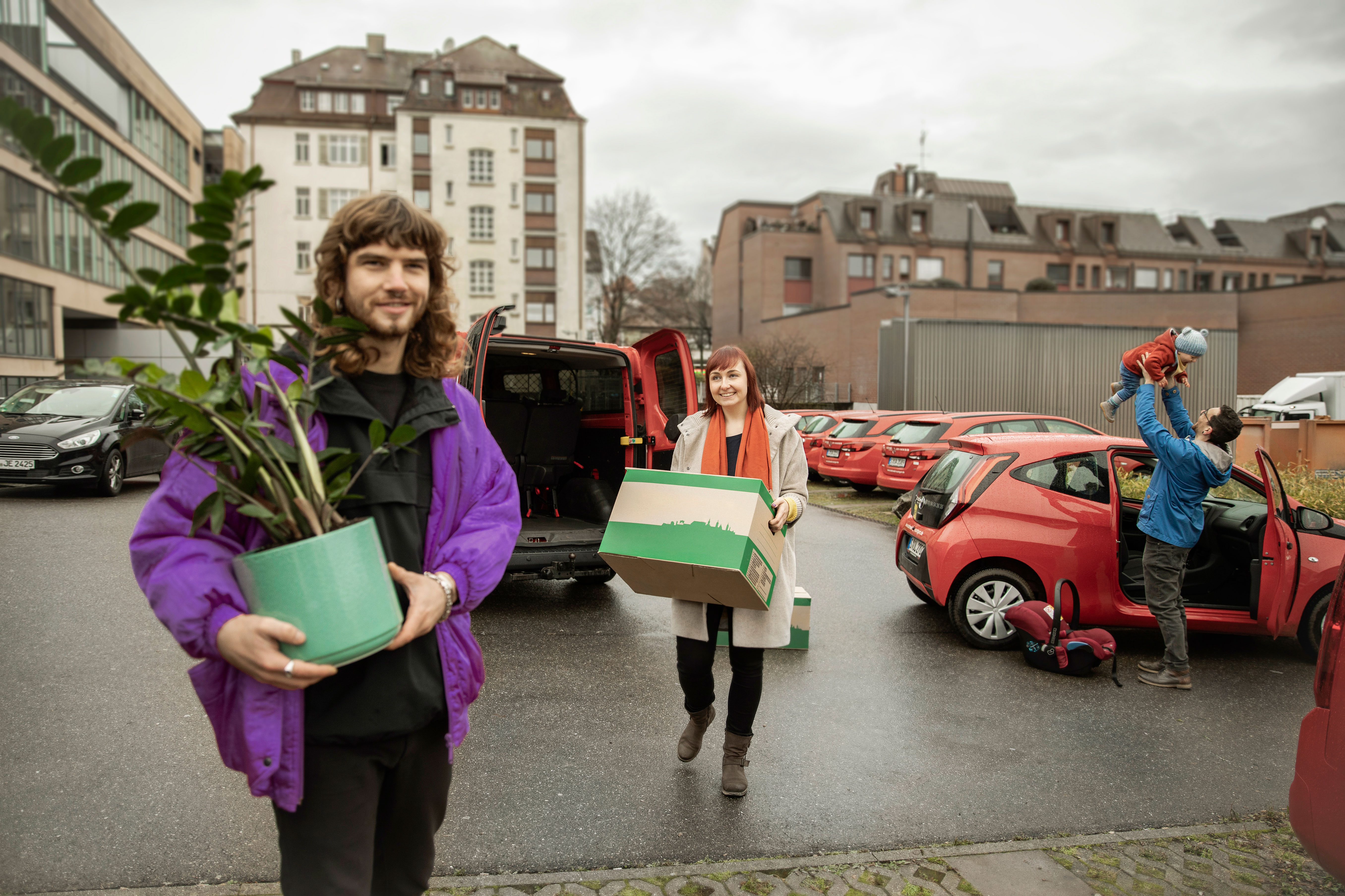 Personen laden Gegenstände aus einem roten Carsharing-Auto, Familie im Hintergrund.