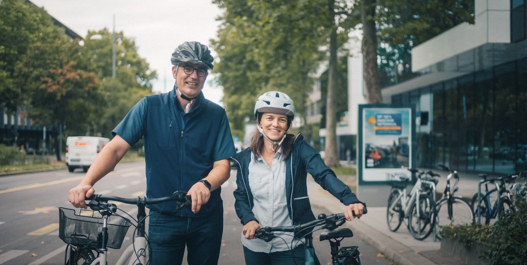 Ein Mann und eine Frau stehen im Freien mit ihren Fahrrädern in der Hand, Helm auf dem Kopf, Straße im Hintergrund.
