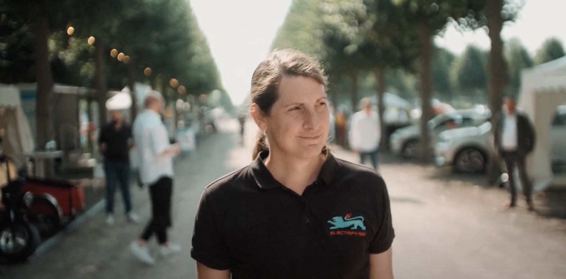 Frau mit schwarzem Polohemd läuft im Freien, Bäume und Personen im Hintergrund.